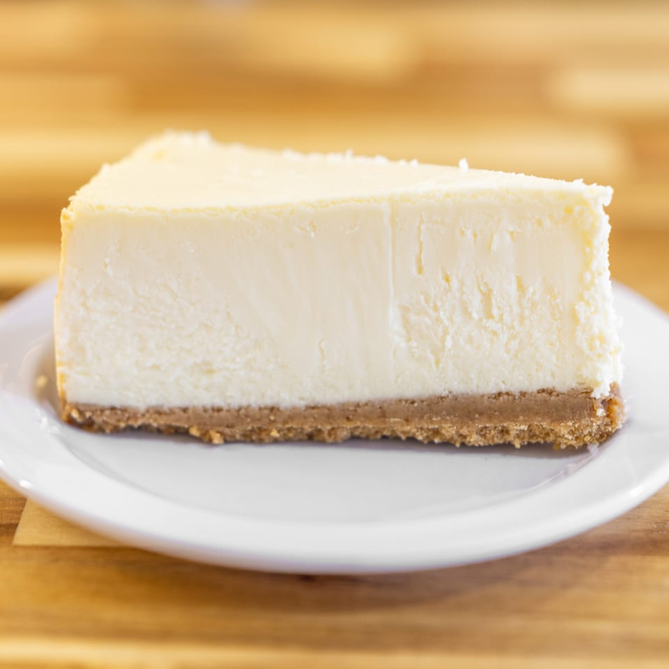 A slice of classic cheesecake with a graham cracker crust on a white plate.