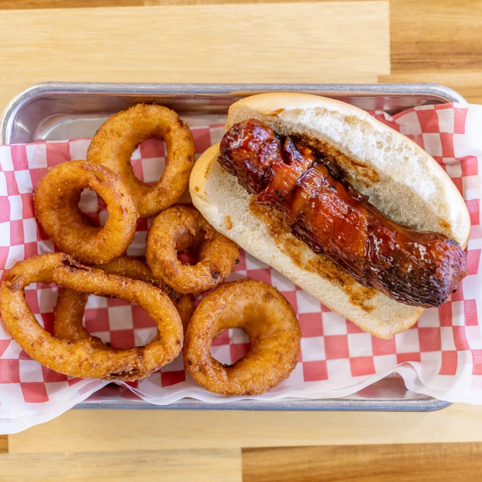 Dave's BBQ & Smokehouse hot dog with BBQ sauce and a side of crispy onion rings.