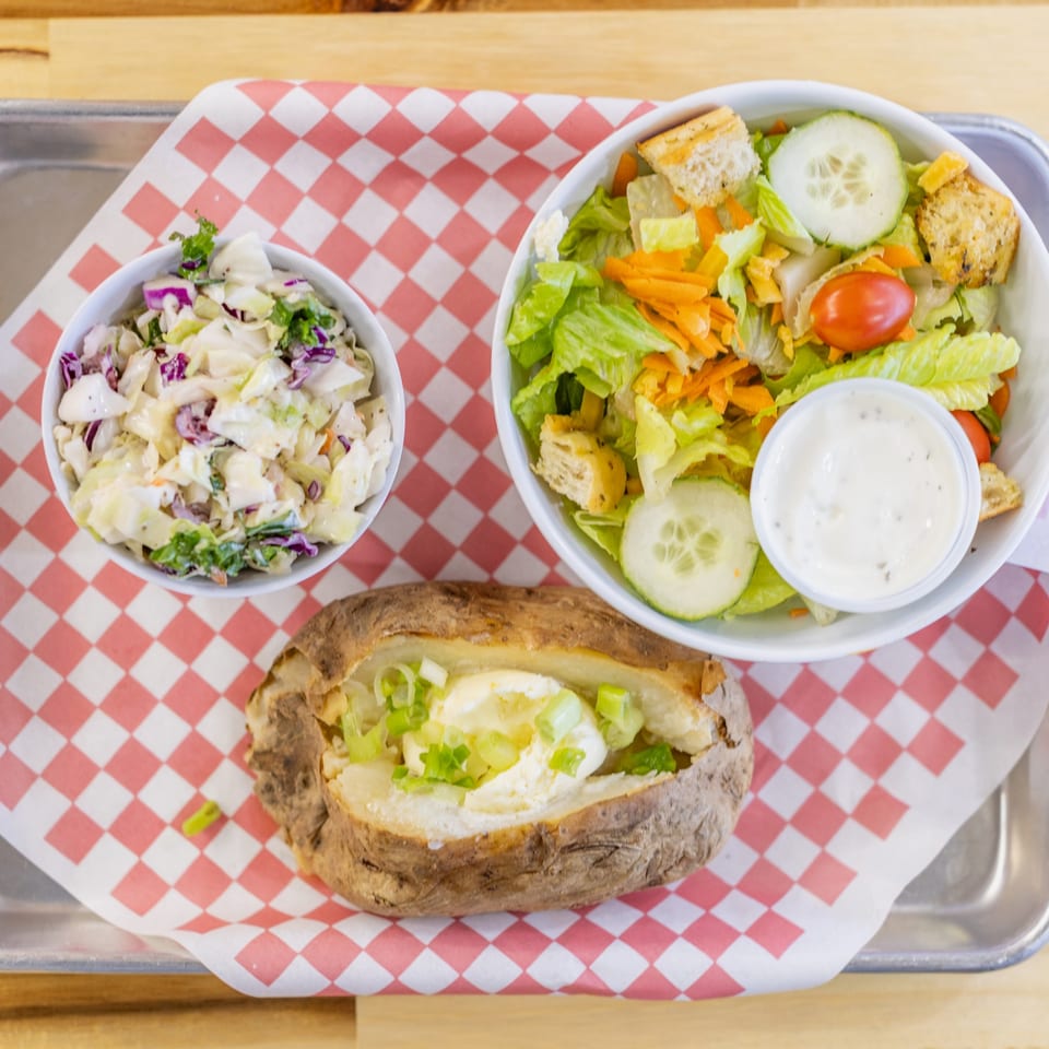 Overhead view of a baked potato with butter and chives, coleslaw, and a side salad with ranch dressing.